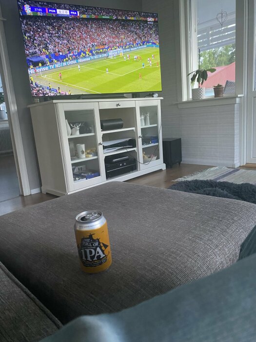 En burk IPA-öl står på en grå ottoman framför en tv som visar en fotbollsmatch mellan Portugal och Frankrike, där matchen står 0-0.