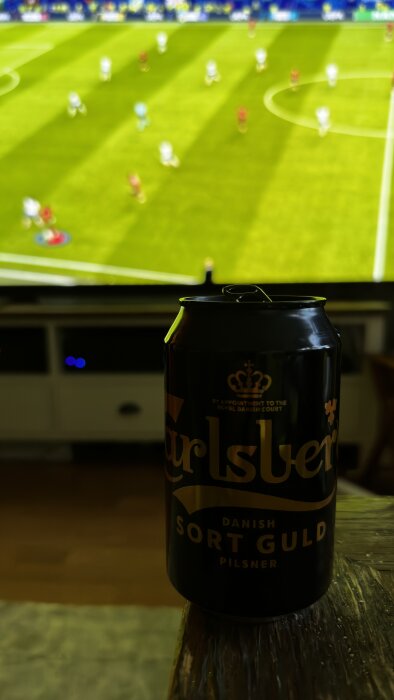 En burk Carlsberg Sort Guld pilsner framför en TV som visar en fotbollsmatch mellan lag med vita och röda tröjor.