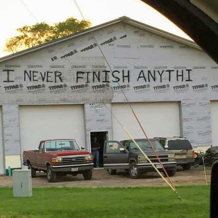 Byggnad med texten "I NEVER FINISH ANYTHI" målad på väggen, omgiven av bilar och en person som står vid en dörr.