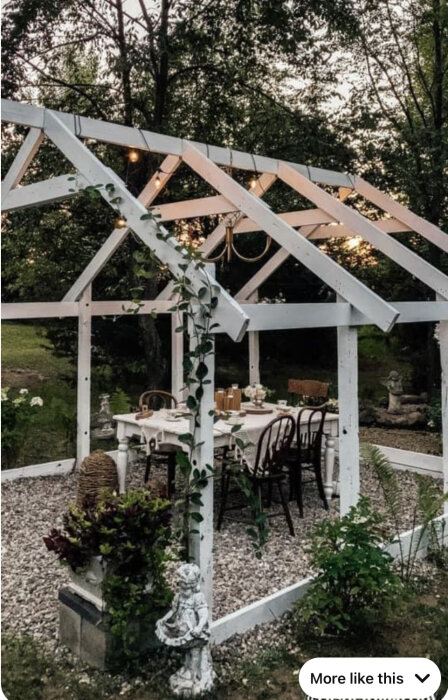 Vit pergola med sadeltak och klätterväxter, inredd med bord och stolar under takbjälkar, omgivet av gröna träd och växter på en grusbelagd yta.