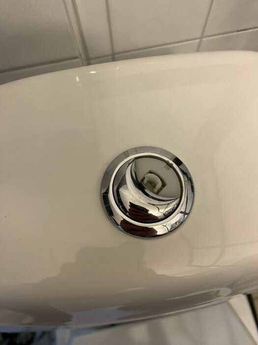 Spolknapp i metall på en vit toalett. Knappen verkar vara trasig och behöver tas bort för att ersättas, vit kakelvägg i bakgrunden.