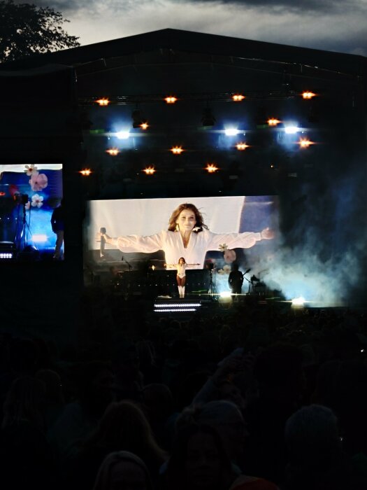 Kvinna på scen med armarna utsträckta under en konsert på kvällen, stora bildskärmar bakom henne och publiken i förgrunden.