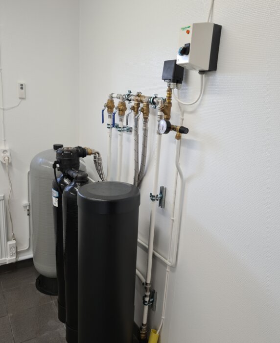Två filtreringsbehållare i en vitmålad teknisk installationsmiljö med synliga röranslutningar och kontrollpanel på väggen.