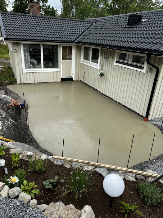 Betonggjuten uteplats framför ett hus med vit fasad och mörkt tak. Betongplattan har ett område som lutar felaktigt och bildar en vattenpöl i mitten.