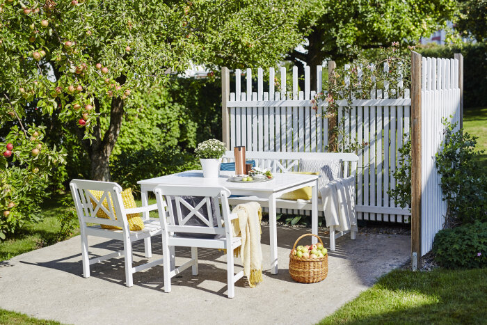 Vit uteplats med träbord, stolar och bänk på cementgolv, omgiven av trädgård med äppelträd. Korg med äpplen och blomkrukor på bordet, vitt staket i bakgrunden.