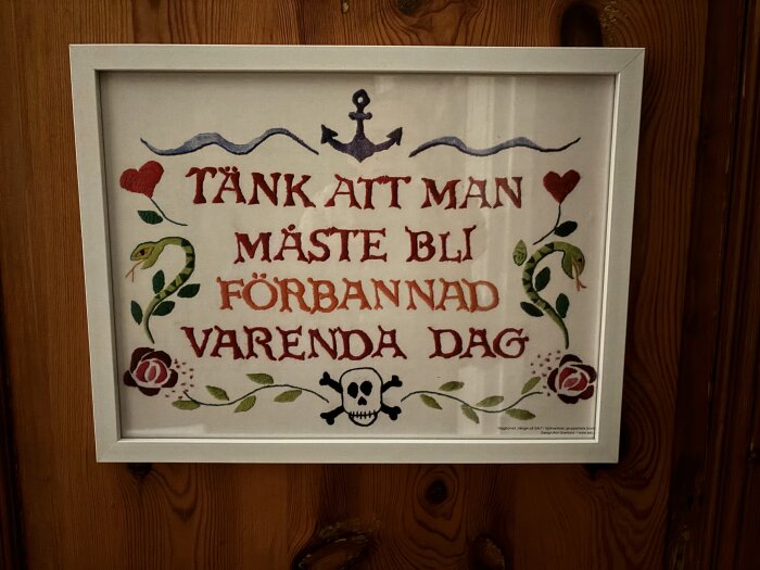 Målning i vit ram med texten "TÄNK ATT MAN MÅSTE BLI FÖRBANNAD VARENDA DAG", pryda av ankare, ormar, hjärtan, blommor och en dödskalle.