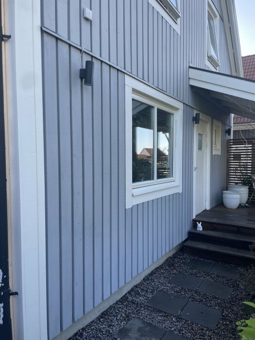 Entrésidan av ett hus med blå stående lockpanel och vita knutbrädor. Trappa och stengång leder till verandadörren.