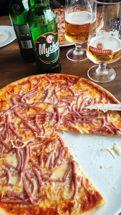 Pizza med skinka, två glas öl märkta med "Mariestads" och flaskor av Mythos-öl på ett träbord.
