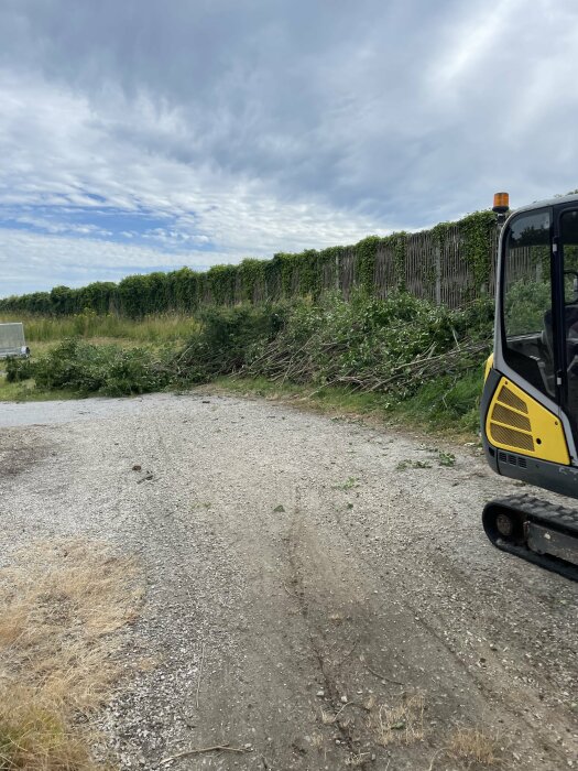 Nytt grusområde efter röjning av buskar med grävmaskin synlig till höger och nedklippt vegetation längs staket i bakgrunden.