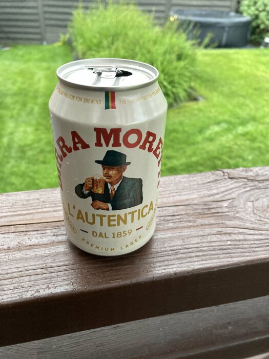 Burk med Birra Moretti L’Autentica öl placerad på ett trästaket i en trädgård med grönt gräs och buskar i bakgrunden.