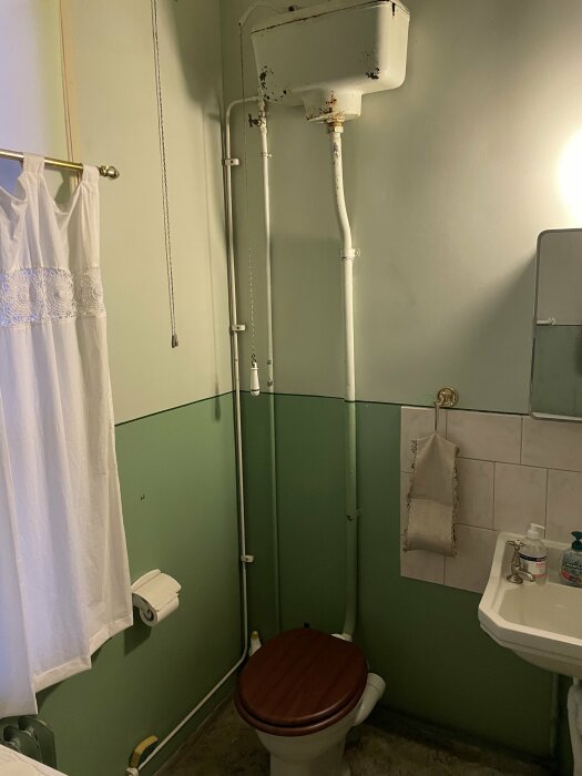 Högspolande toalett med tank placerad ovanför toaletten i ett fritidshusbadrum. Tanken har ingående vattenrör från höger sida och en kedja att dra i för att spola.