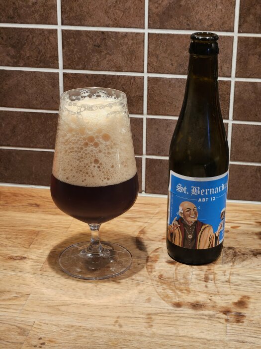 Ett glas med mörk öl och ett ölflaska av märket "St. Bernardus Abt 12" på ett träbord framför en brun kaklad vägg.