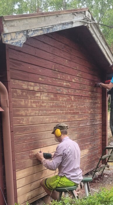 Man slipar vägg av röd träbyggnad med en slipmaskin; andra person målar takutsprång på byggställning.