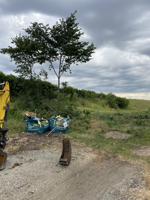 Tre stora säckar stående i grönt område bredvid en skopa under träd och molnig himmel. Grusig yta i förgrunden.