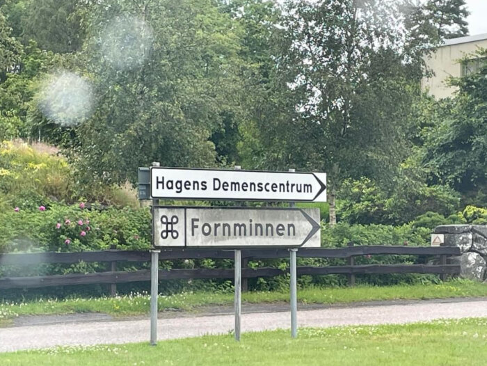 Vägskyltar vid en landsväg, en pekar mot "Hagens Demenscentrum" och en annan mot "Fornminnen", omgivna av lummig grönska och blommor.