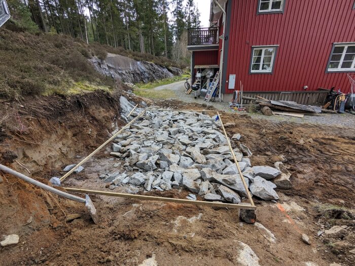 Grundläggning med grova granitstenar inom en avgränsning; omgivande jord och skog i bakgrunden. Röd träbyggnad syns till höger i bilden.