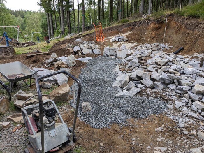 En grundmur i granit under uppbyggnad med sprängstenar och dräneringsmaterial, omgiven av ett jordigt område med grön vegetation och byggverktyg i förgrunden.