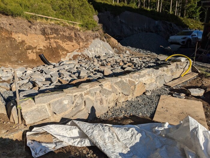 Grundmur i granit med oregelbundna stenar, makadam, och dräneringsrör på en byggarbetsplats. Duk och träskivor ligger framför muren.