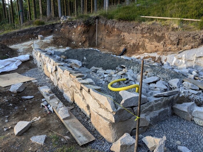 Byggarbetsplats med en grundmur av oregelbundna granitstenar under konstruktion, omgiven av jord, grus och byggmaterial.