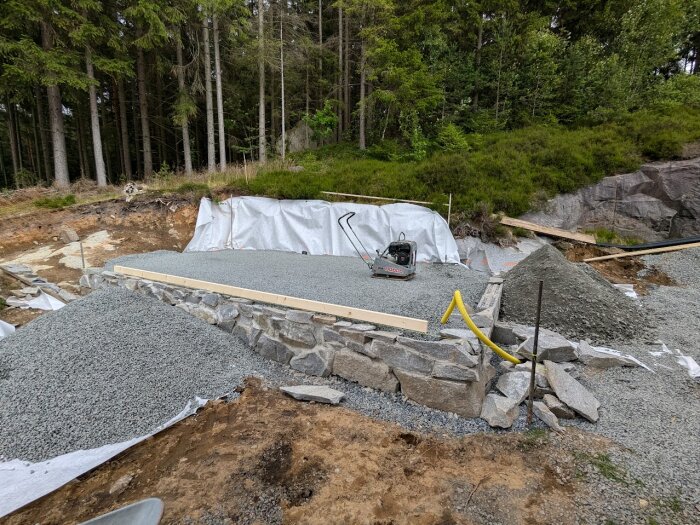 En grundmur i granit under uppbyggnad med en staplad stenmur, gruslager och verktyg i en skogsmiljö.