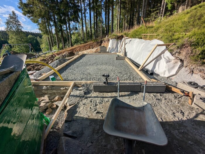 Formarbete och armering för en ~10cm tjock betongplatta med 45x95 reglar, grusgrund, stödmur och skottkärra i förgrunden, omgiven av skog.