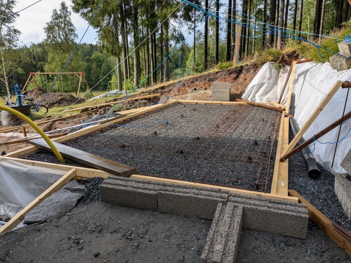 En byggarbetsplats med en betongform gjord av träreglar, armeringsjärn och nät förbereder för gjutning. Omkringliggande träd och material syns i bakgrunden.