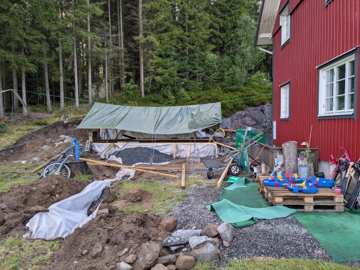 Byggarbetsplats med pågående formbygge och armering för betongplatta, omgiven av skog och intill ett rött hus. Presenning skyddar området från regn.