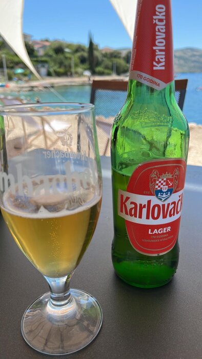 Flaska med Karlovačko lager och ett glas öl på ett bord med en strand och hav i bakgrunden.
