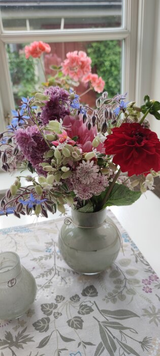 En bukett med blandade blommor i olika färger och former i en grönaktig vas, stående på en blommig duk med fönsterbakgrund.