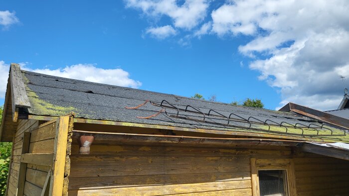 Slitet papptak på gammalt garage med mossväxt, söndriga takpannor och skorstensrester. Blå himmel med moln i bakgrunden.