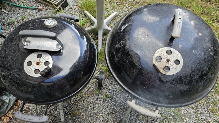 Två runda Weber-grillar med lock ståendes på grus, en med termometer och den andra utan. Trädgårdsslang och växtlighet i bakgrunden.