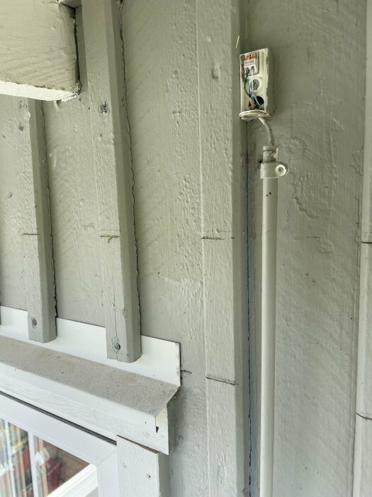 Vit elektroteknisk givare monterad på grön husvägg, med kabel som leder neråt mot pannrummet i källaren, ovanför ett fönster.