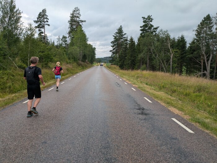 Två personer som springer på en landsväg omgiven av skog och gräs med en bil synlig i fjärran.