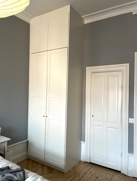 Vit garderob placerad mellan en vägg och en rundad pelare i ett rum med grå väggar och parkettgolv. En dörr syns till höger om garderoben.