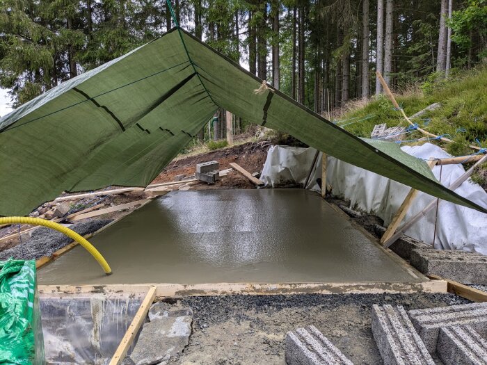 Nygjuten betongplatta täckt av en grön presenning, omgiven av betongblock och byggmaterial, med en tallskog i bakgrunden.