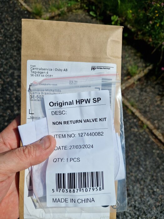 En person håller i ett paket med etiketten "Original HPW SP", en backventilsats. Paketet är från Centralservice i Osby AB och ankomstberäknat till 27/03/2024.