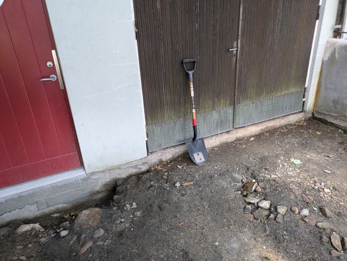 Grävd grop framför husvägg med rödfärgad dörr och intilliggande garageport. En spade står lutad mot väggen.
