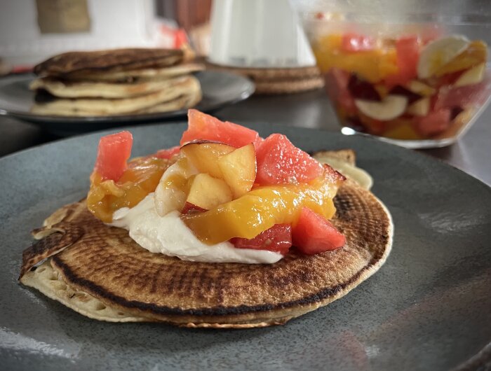 Amerikanska pannkakor toppade med fruktsallad och vispad grädde på en tallrik, fruktsallad i en skål i bakgrunden.