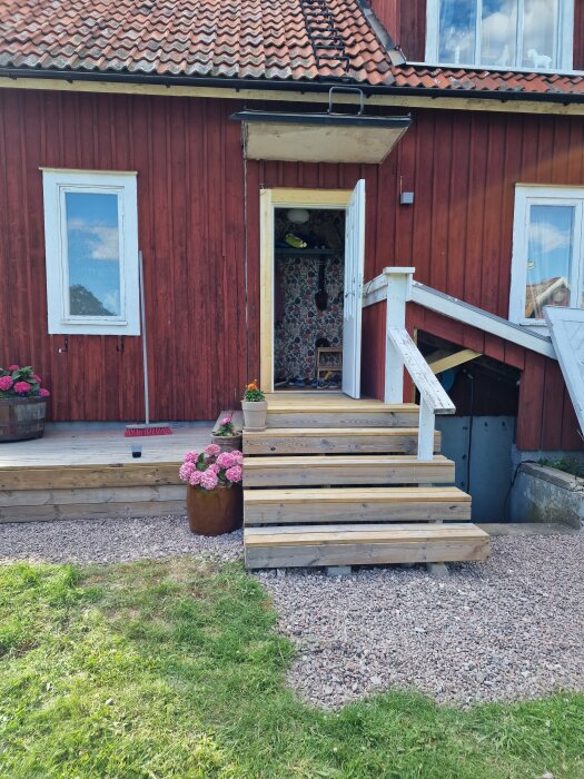 Tillfällig entré i träd med tillhörande trappa framför ett rött hus och två krukor med blommor. Skiffergång och trädäck synliga.