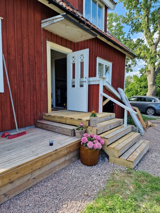 Tillfällig entré med nya och begagnade plankor, trappor i flera nivåer, blomkrukor med rosa och orange blommor, vita dörrar och rött hus i bakgrunden.