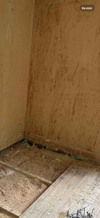 Brunt rumshörn med synlig isoleringsvadd. Gröna fibrer sträcker sig ur nedre delen av väggen till golvet där sågspån och spik ligger utspridda.