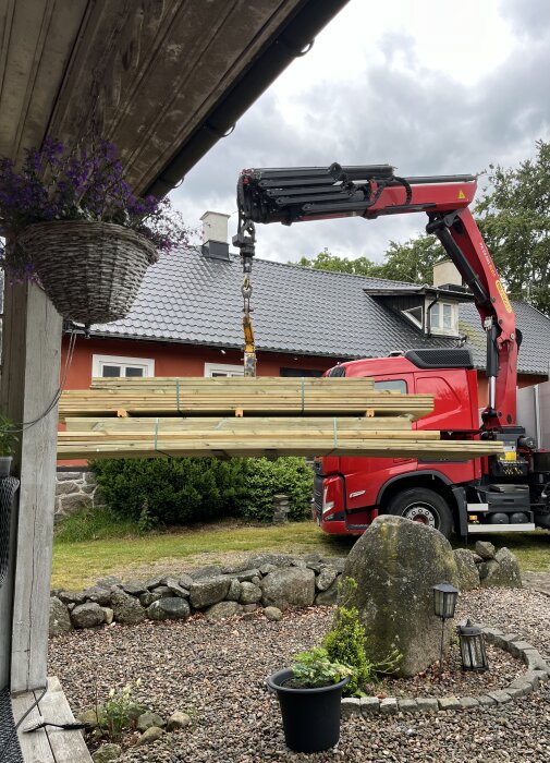 Kranbil lossar en stapel virke vid ett hus med träpanel och tegeltak, stenbelagd uppfart och en blomkruka i förgrunden.