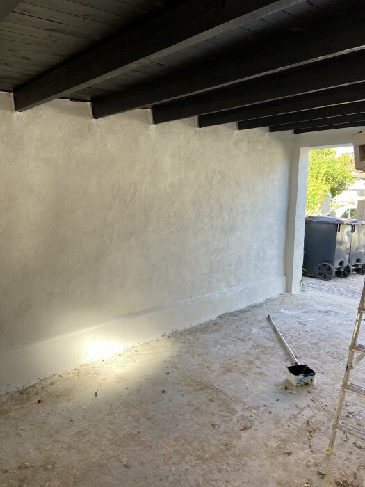 Garagetak med synliga bjälkar som sluttar nedåt mot en nyputsad vägg. På golvet ligger en målarroller bredvid sin färgbehållare och en stege står vid väggen.