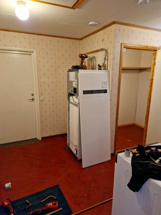 Tvättstuga med röd matta, vit värmepump mitt i rummet, avstängd i ena hörnet. Inkommande rör synliga, verktyg och delar utspridda på golvet.