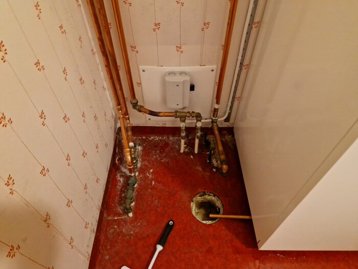 Tvättstuga med synliga kopparrör, en radiator och ett hål i det röda golvet; vägghängd enhet för värmepump på mönstrad tapet.