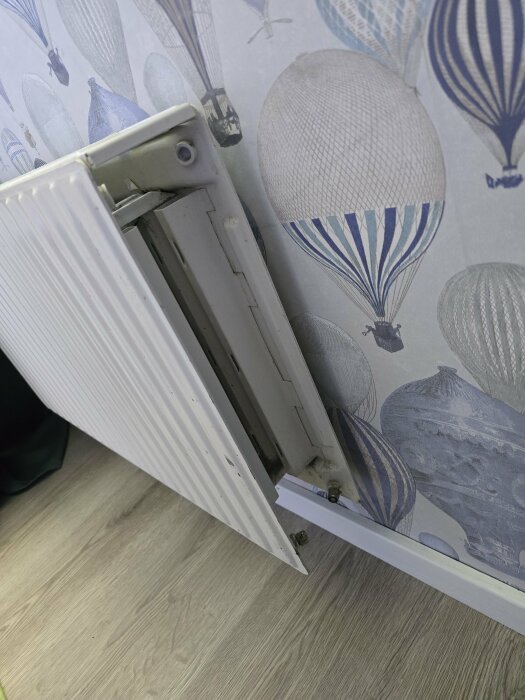 Öppen radiator vid vägg med luftballongstapet, underdelen visar rör och skruvar. Golvet är i träimitation.