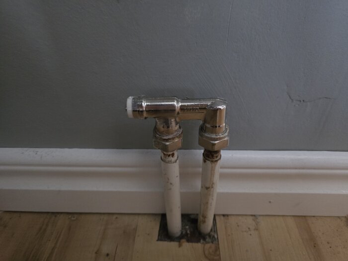 Två vita rör med metallkopplingar vid golvet och väggen i en tvättstuga.