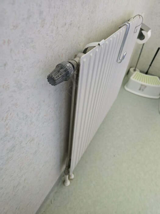 En vit radiator monterad på en vägg i ett rum. Radiatorn har en termostatventil märkt med siffror och rör som går ner till golvet.