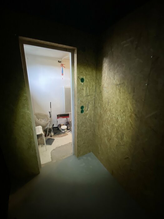 Renoverad vägg med OSB-skivor och en öppning som leder till ett rum med byggmaterial, inklusive en målarburk och stege, endast upplyst av pannlampa.