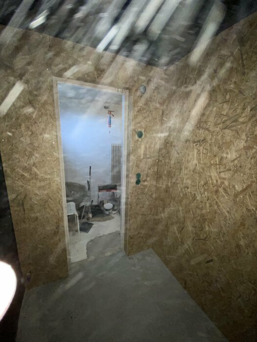 Bild av ett rum under renovering med osb-skivor på väggarna och ett öppet dörrhål till ett annat rum med byggrör och stolar.